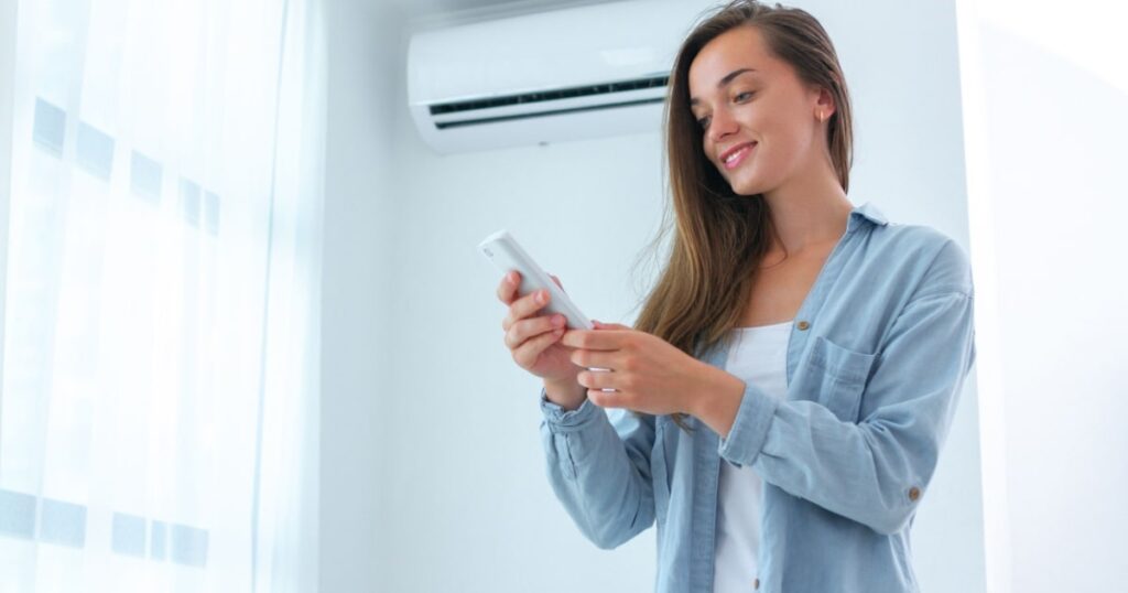 Une jeune femme dos à une unité intérieure de climatisation en tenant une télécommande. 