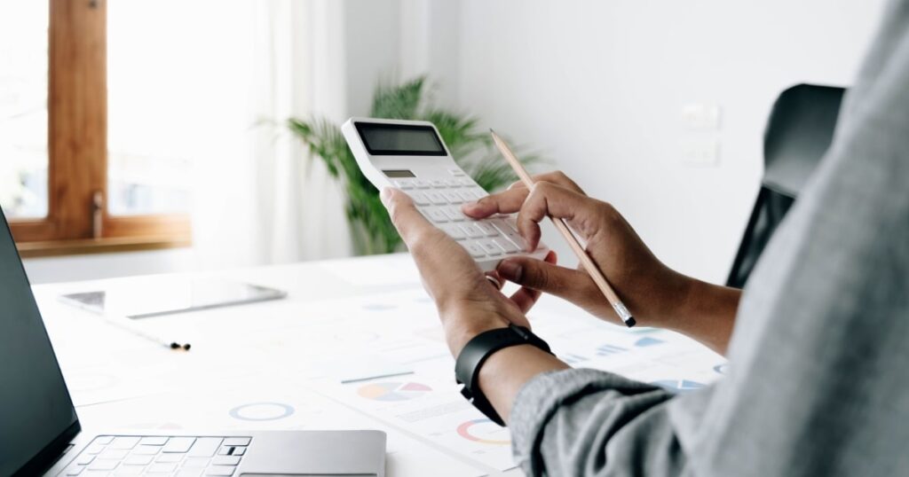 Les mains d'un homme assis à une table devant un ordinateur tapent sur une calculatrice.